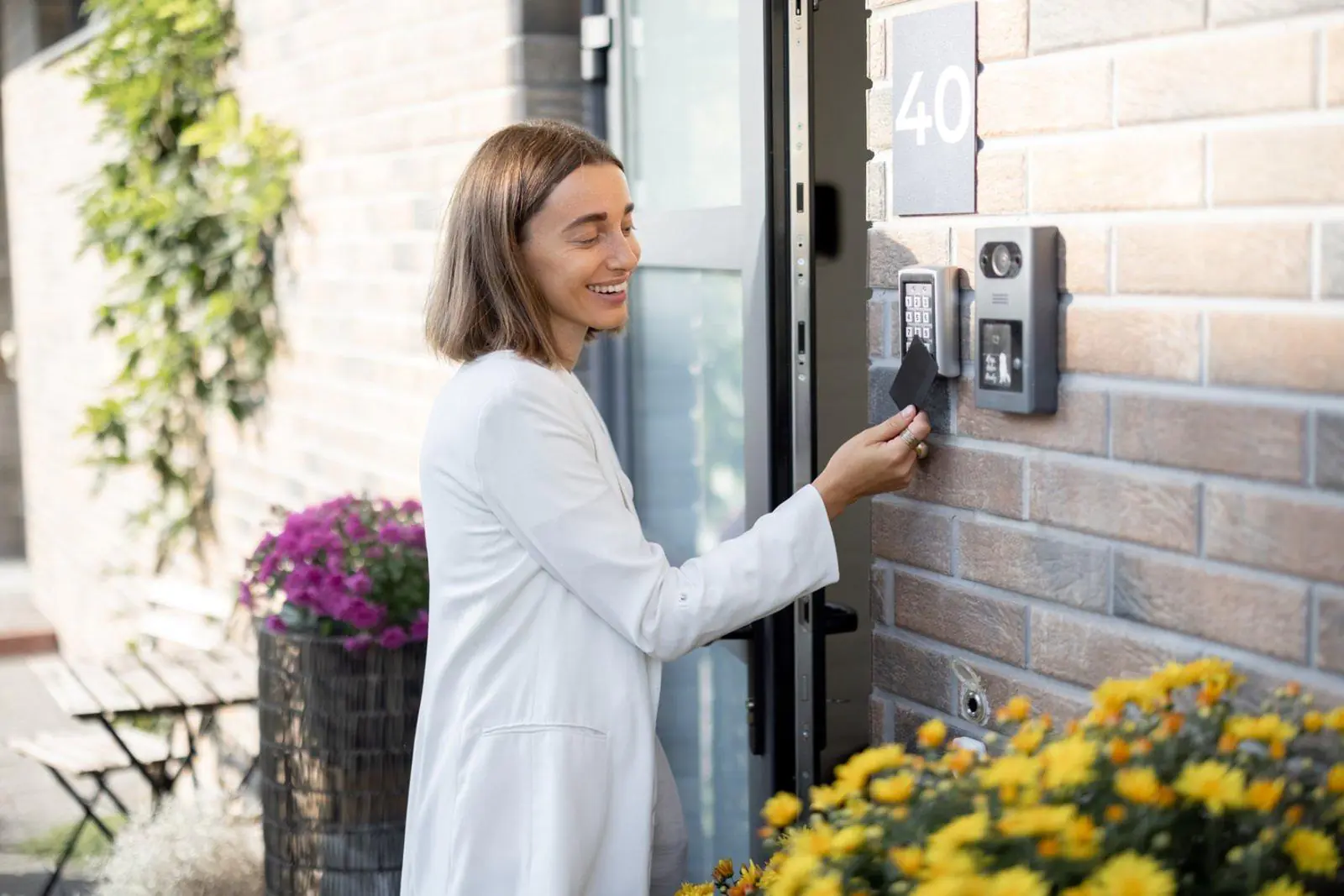 A woman in a white blazer smiling while using a keycard on a digital intercom system at the entrance of a building, representing the convenience of modern Virtual Doorman Services
