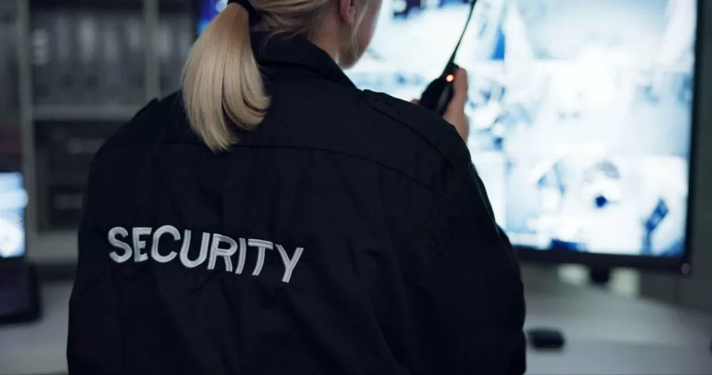 Rear view of a security guard with blonde hair, wearing a black "SECURITY" jacket, holding a two-way radio and looking at monitoring screens as part of a CCTV Service