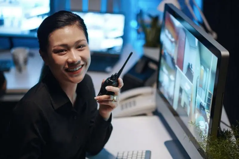 A female Virtual Doorman with a walkie-talkie smiles while monitoring security camera feeds on her computer, demonstrating the duties and responsibilities of modern doorman services