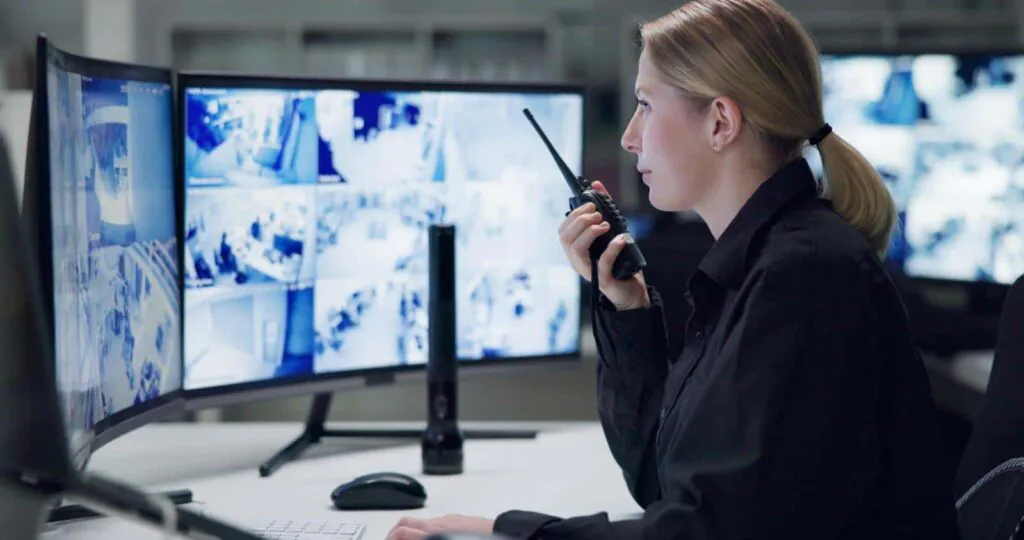 A professional female security operator in a black uniform uses a radio while monitoring multiple surveillance screens, illustrating the human expertise that influences Camera Monitoring Service Cost