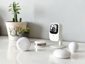 A collection of white smart home security devices, including a modern Nanny Cam, smoke sensor, and flood sensor, arranged on a white table next to a potted cactus to illustrate Nanny Cam Services