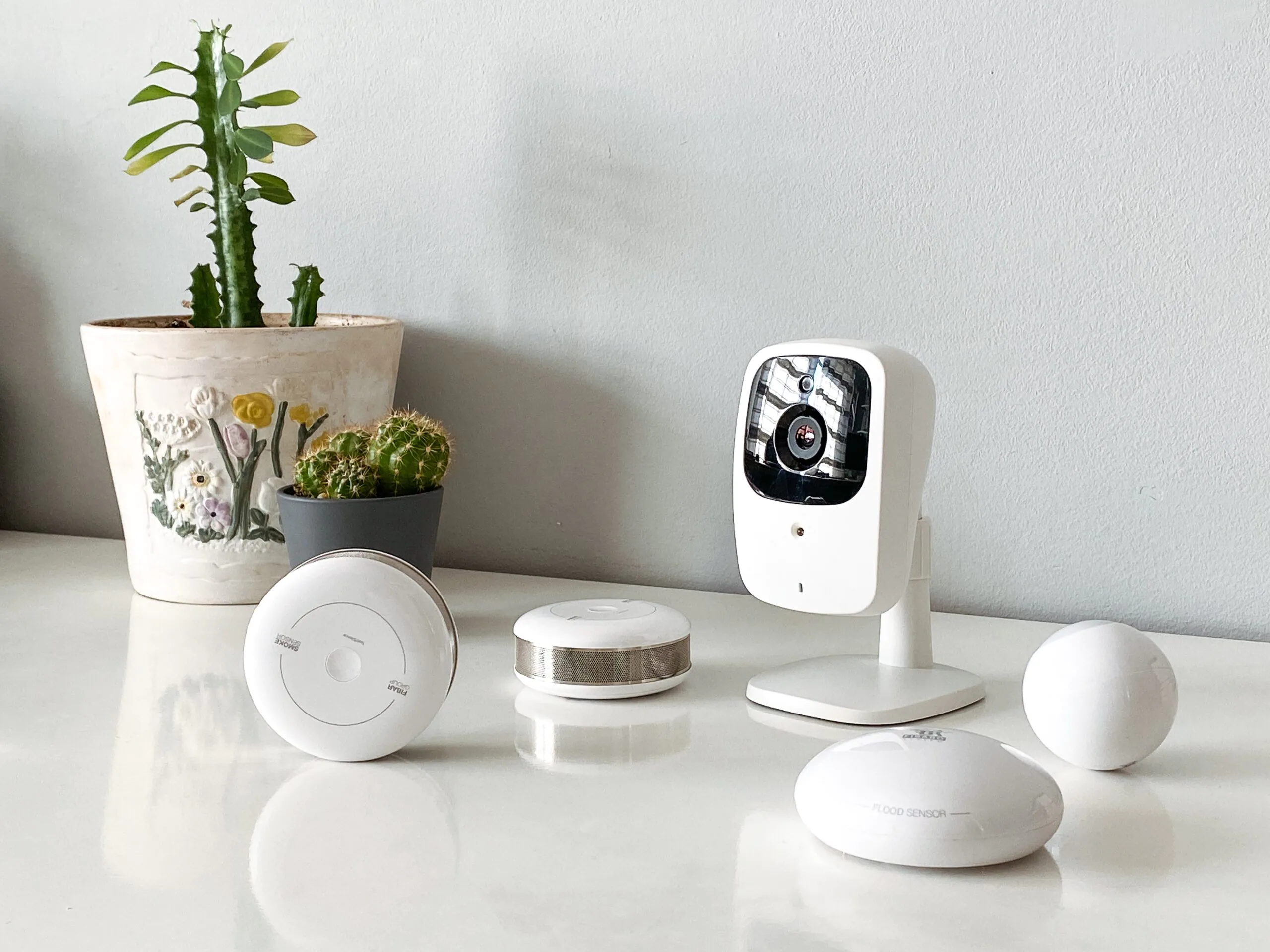 A collection of white smart home security devices, including a modern Nanny Cam, smoke sensor, and flood sensor, arranged on a white table next to a potted cactus to illustrate Nanny Cam Services