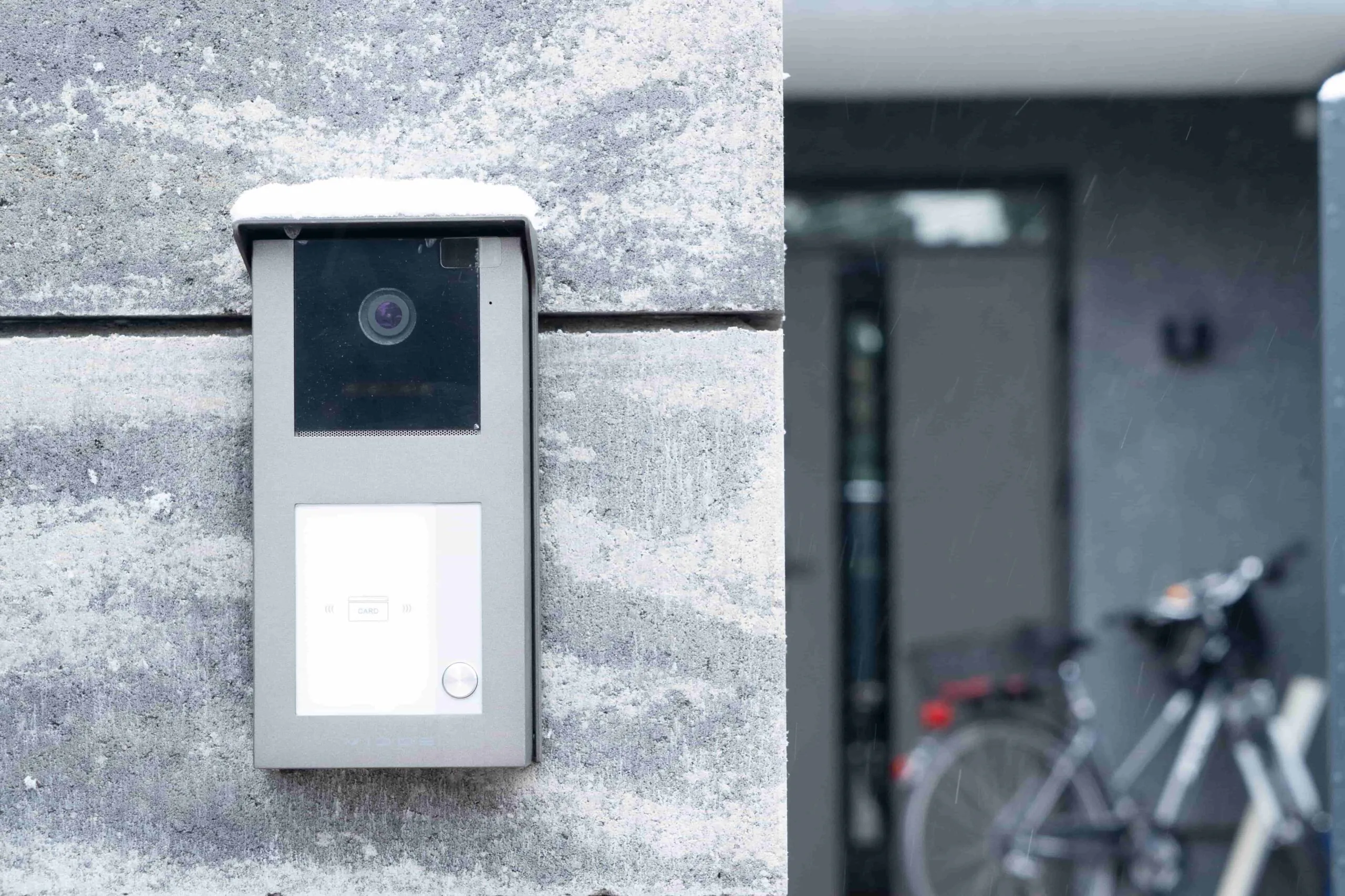 A close-up of a sleek video intercom system mounted on a snowy stone wall, functioning as a high-tech Apartment Doorman for enhanced Building Security at a modern Apartment.