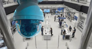 A close-up photograph of a blue dome CCTV Monitoring for Retail Stores security camera mounted high in a busy electronics store, actively overseeing customers and staff visible in the blurred background to illustrate retail security and shoplifting prevention functions.