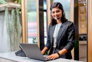 A smiling professional woman wearing a headset and working on a laptop at a modern front desk, perfectly illustrating the Remote Concierge Basics provided by a dedicated Concierge Company serving as a Virtual Receptionist.