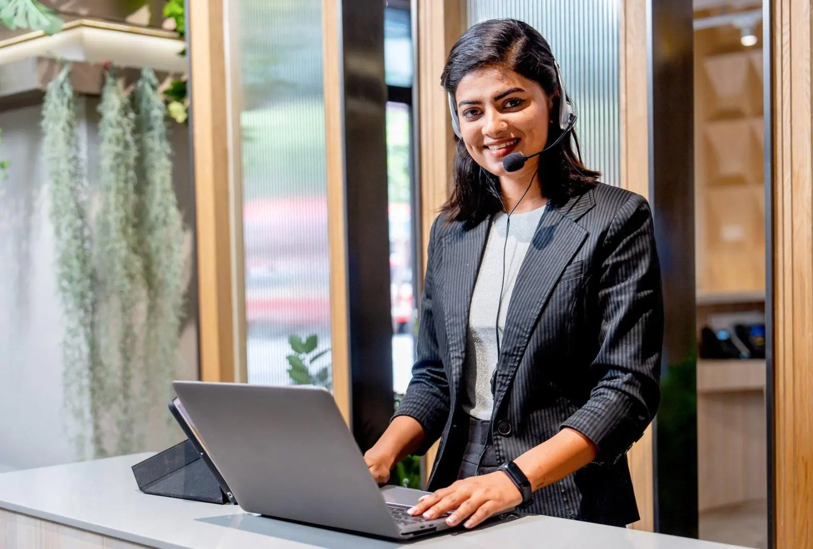 A smiling professional woman wearing a headset and working on a laptop at a modern front desk, perfectly illustrating the Remote Concierge Basics provided by a dedicated Concierge Company serving as a Virtual Receptionist.