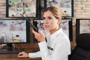 What Apartment Doorman Do: A smiling security professional, holding a communication radio at a computer desk, monitors multiple screens displaying live camera feeds from residential units, managing access control in modern doorman apartments or a traditional NYC doorman building. This scene (image_6.png) illustrates both the physical 'duties of a doorman' and advanced technology like remote doorman and virtual doorman services.