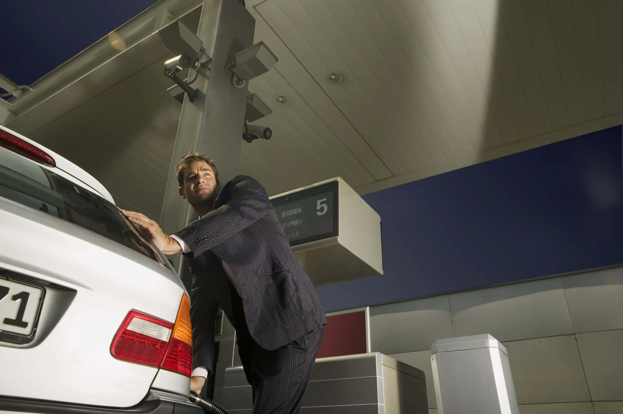 A cautious man in a suit pumps fuel into a silver car while looking over his shoulder, under the watchful eye of multiple overhead security cameras, highlighting the critical importance of CCTV Monitoring for Gas Stations for effective surveillance monitoring and security measure monitoring.
