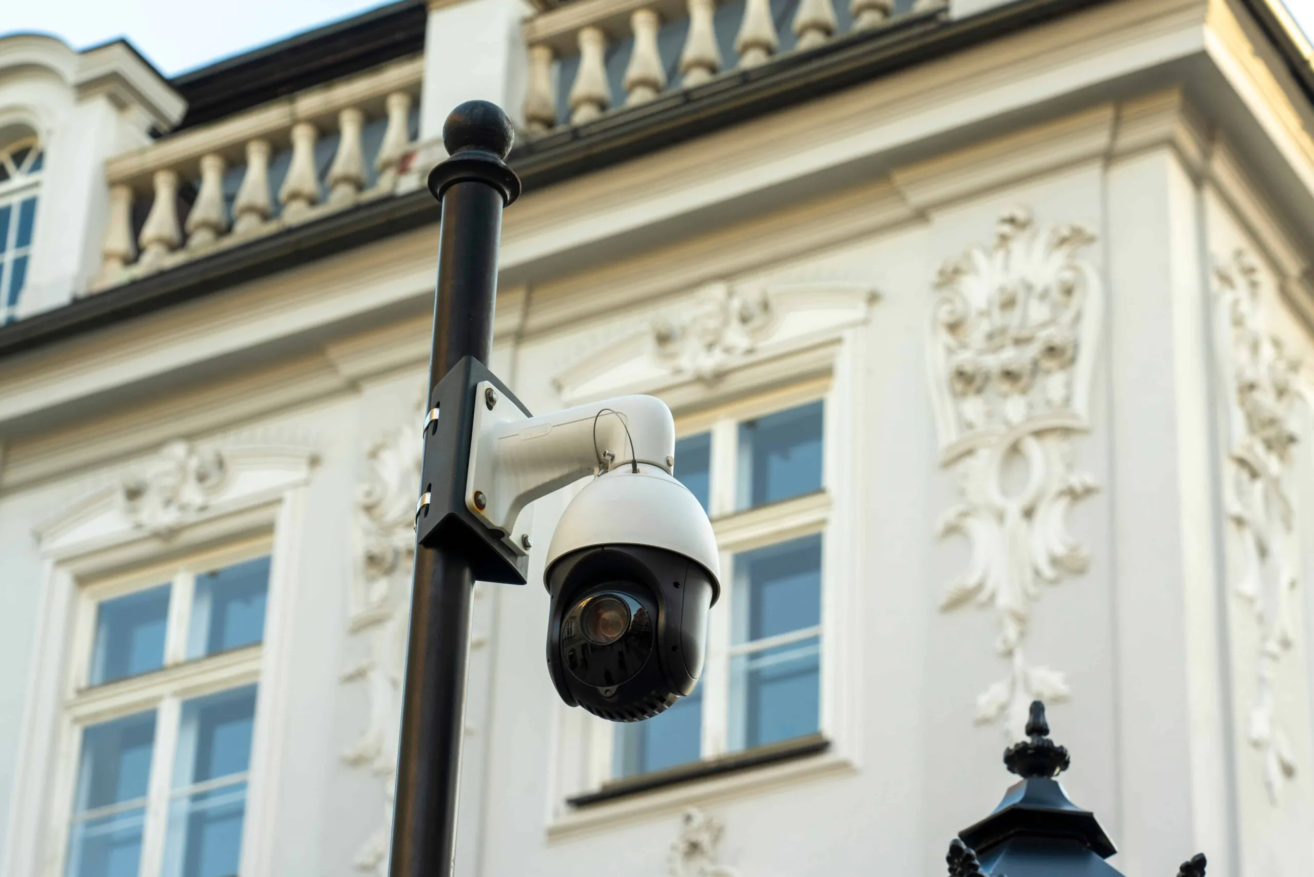 Alt Text: A high-tech PTZ dome security camera mounted on a black pole outside an elegant, ornate white building, illustrating the robust hardware required in CCTV Monitoring For Hotels for complete hotel security and continuous surveillance monitoring.