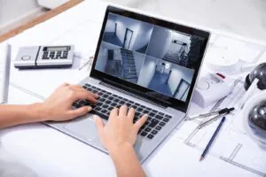 A person evaluating if CCTV monitoring worth it by reviewing a live security camera monitoring feed on a laptop surrounded by architectural blueprints and video surveillance hardware.