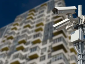 A pair of outdoor bullet security cameras securely mounted on a metal pole in front of a multi-story residential building, highlighting the importance of CCTV monitoring for apartments to ensure comprehensive apartment security and effective building security through a reliable CCTV monitoring service.