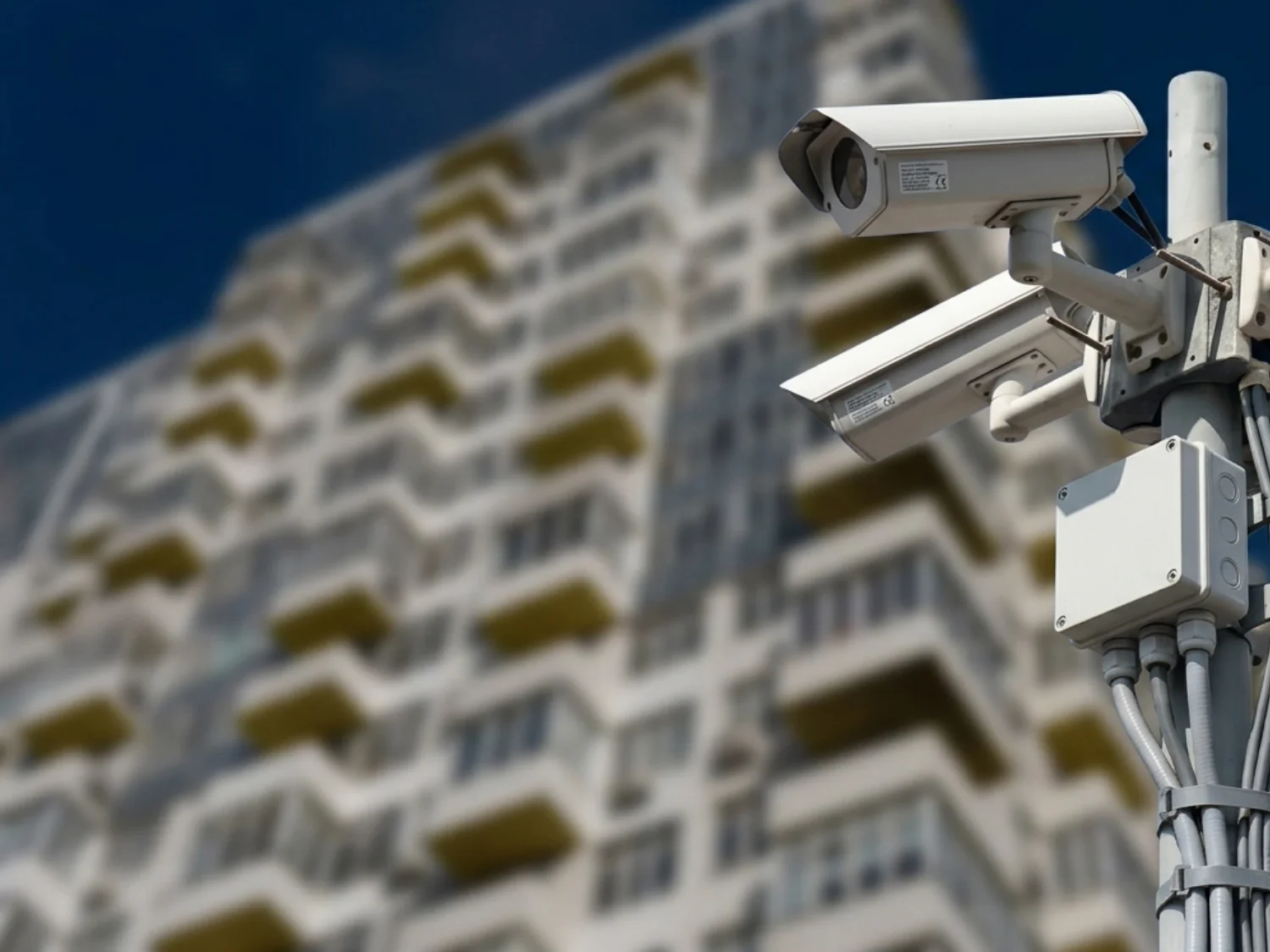 A pair of outdoor bullet security cameras securely mounted on a metal pole in front of a multi-story residential building, highlighting the importance of CCTV monitoring for apartments to ensure comprehensive apartment security and effective building security through a reliable CCTV monitoring service.