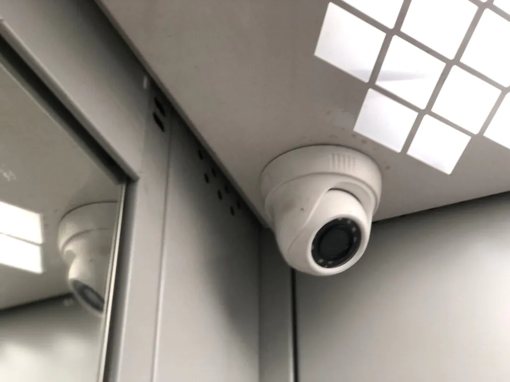 A close-up of a white dome security camera mounted in the corner of a corporate elevator ceiling next to a mirrored wall, demonstrating the strategic placement required for effective CCTV monitoring for office environments by a reliable CCTV monitoring services provider to ensure complete security surveillance.