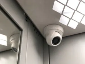 A close-up of a white dome security camera mounted in the corner of a corporate elevator ceiling next to a mirrored wall, demonstrating the strategic placement required for effective CCTV monitoring for office environments by a reliable CCTV monitoring services provider to ensure complete security surveillance.
