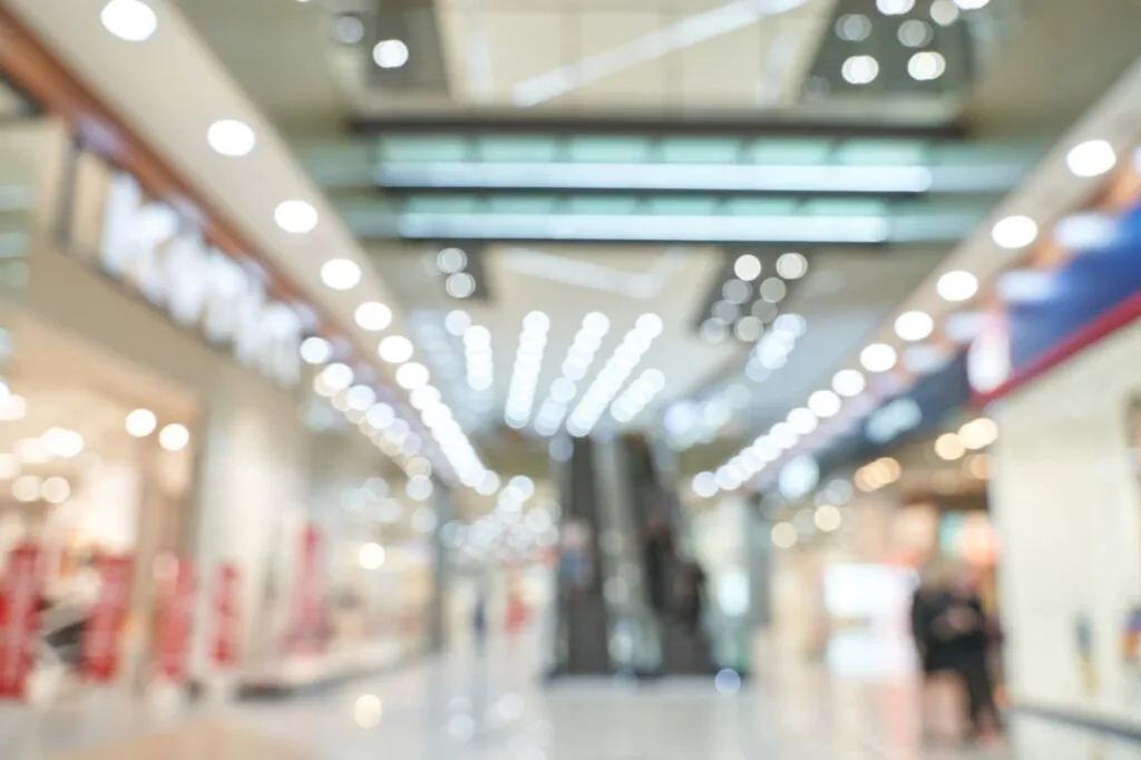 A blurred background view of a brightly lit interior of a shopping center with escalators, representing the expansive public areas that require comprehensive CCTV Monitoring for Shopping Malls using a reliable cctv monitoring service and commercial security cameras to ensure optimal shopping center security.