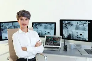 A professional female security specialist with arms crossed sitting in front of multiple monitors displaying split-screen multi-camera feeds, illustrating the operational setup of advanced business cctv systems.
